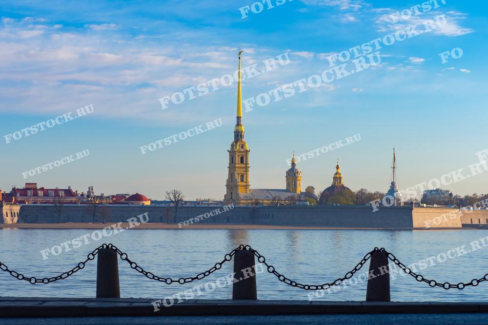 На фотографии изображена Петропавловская крепость, расположенная на берегу Невы в Санкт-Петербурге. Величественные башни и золотой шпиль собора создают впечатляющий вид на фоне утреннего неба. Спокойная водная гладь отражает крепость, добавляя снимку глубины и красоты. Этот вид подчеркивает историческую значимость и архитектурное великолепие Петербурга.
